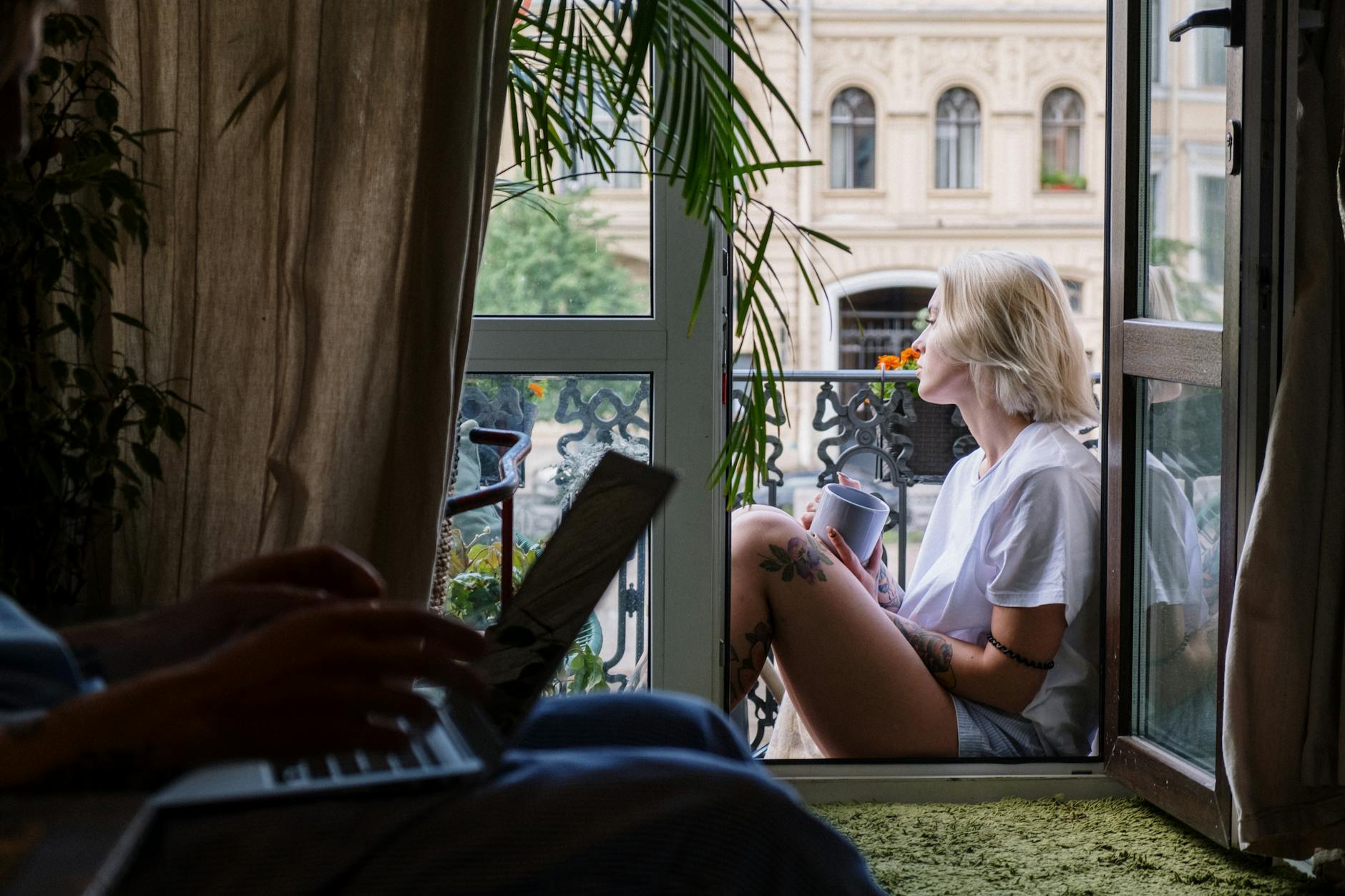 Mały balkon jako letnie biuro: jakie meble i dodatki wybrać, by pracować wygodnie Kobieta z kawą odpoczywa na małym balkonie, laptop stoi w pokoju